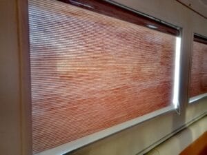 Window with closed brown horizontal blinds, beige window frame, slight light seeping through at the right side.