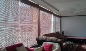 Sunlit lounge with large horizontal blinds casting striped shadows on brown and white seating; a round coffee table sits between cushions, and a white wall or projector screen is visible on the right.