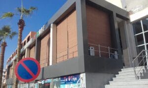 No parking sign in the foreground of a modern storefront plaza with palm trees and blue sky behind it is visible outside a gray and brown building with large windows and shutters.