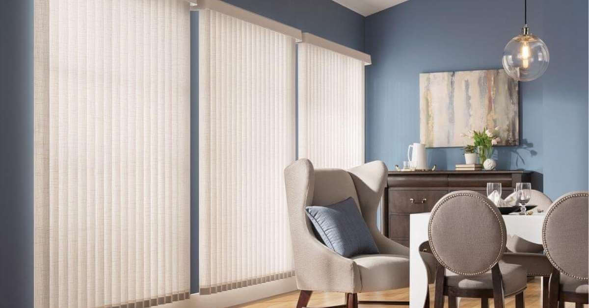 Bright dining area with blue walls, vertical blinds, and beige upholstered chairs around a white table set with glassware.