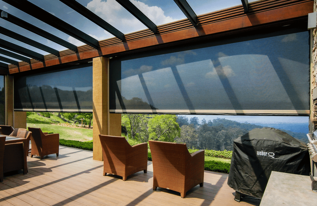 Covered outdoor patio with wicker chairs, overlooking a green hillside; a Weber grill is covered on the right side under a glass pergola.