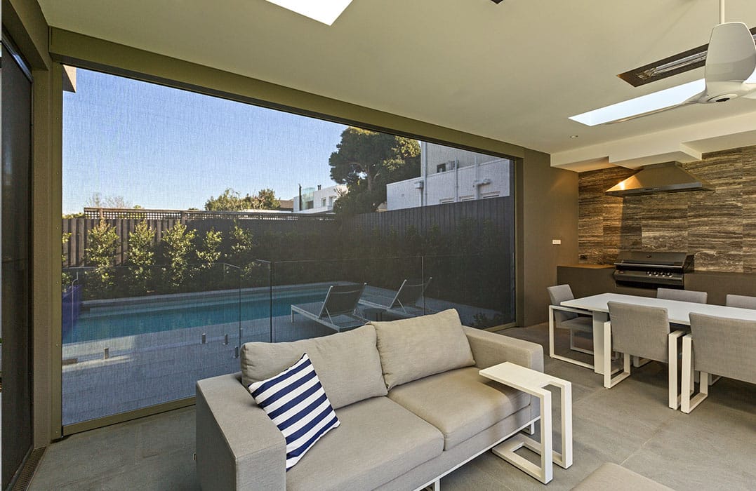 Contemporary indoor–outdoor living space with a glass wall overlooking a pool, beige sofa with a striped pillow, and a white dining table area with a built‑in grill nearby.