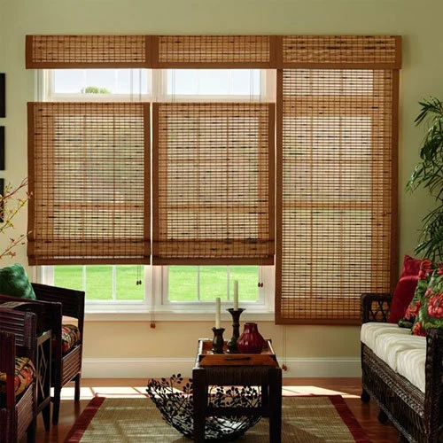 Living room with large windows covered by woven bamboo blinds, letting warm light through between wicker chairs and a coffee table with candles.