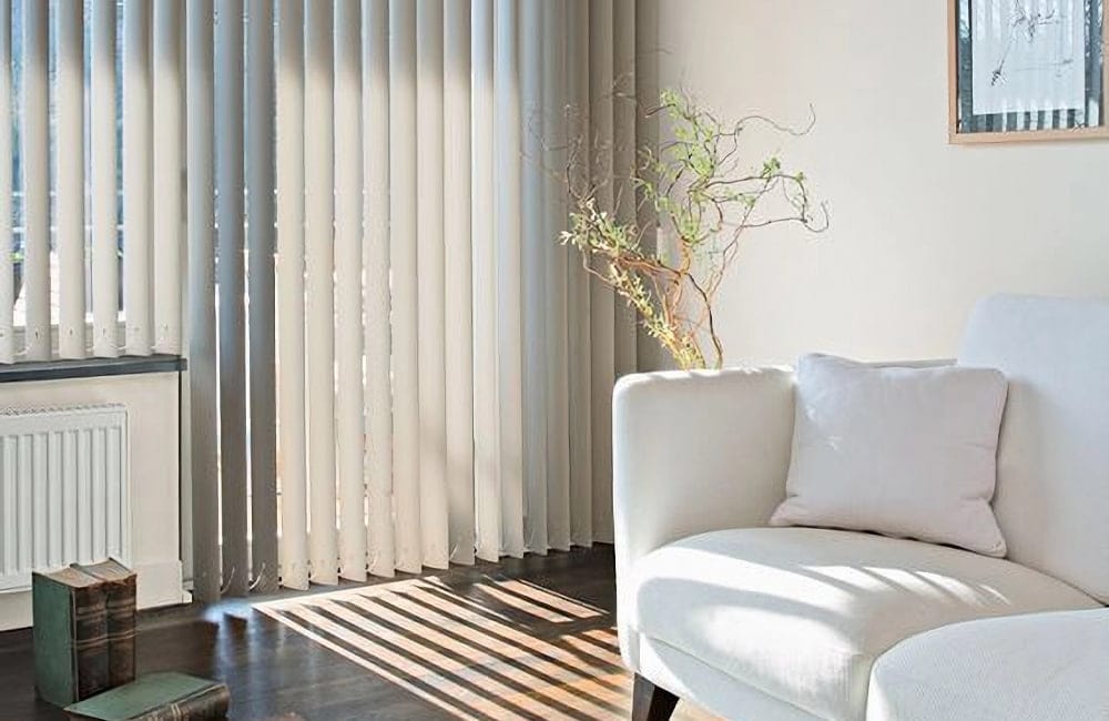 Bright living room corner with a white sofa, pale pillow, tall vertical blinds, and a bare-branch plant beside a radiator.