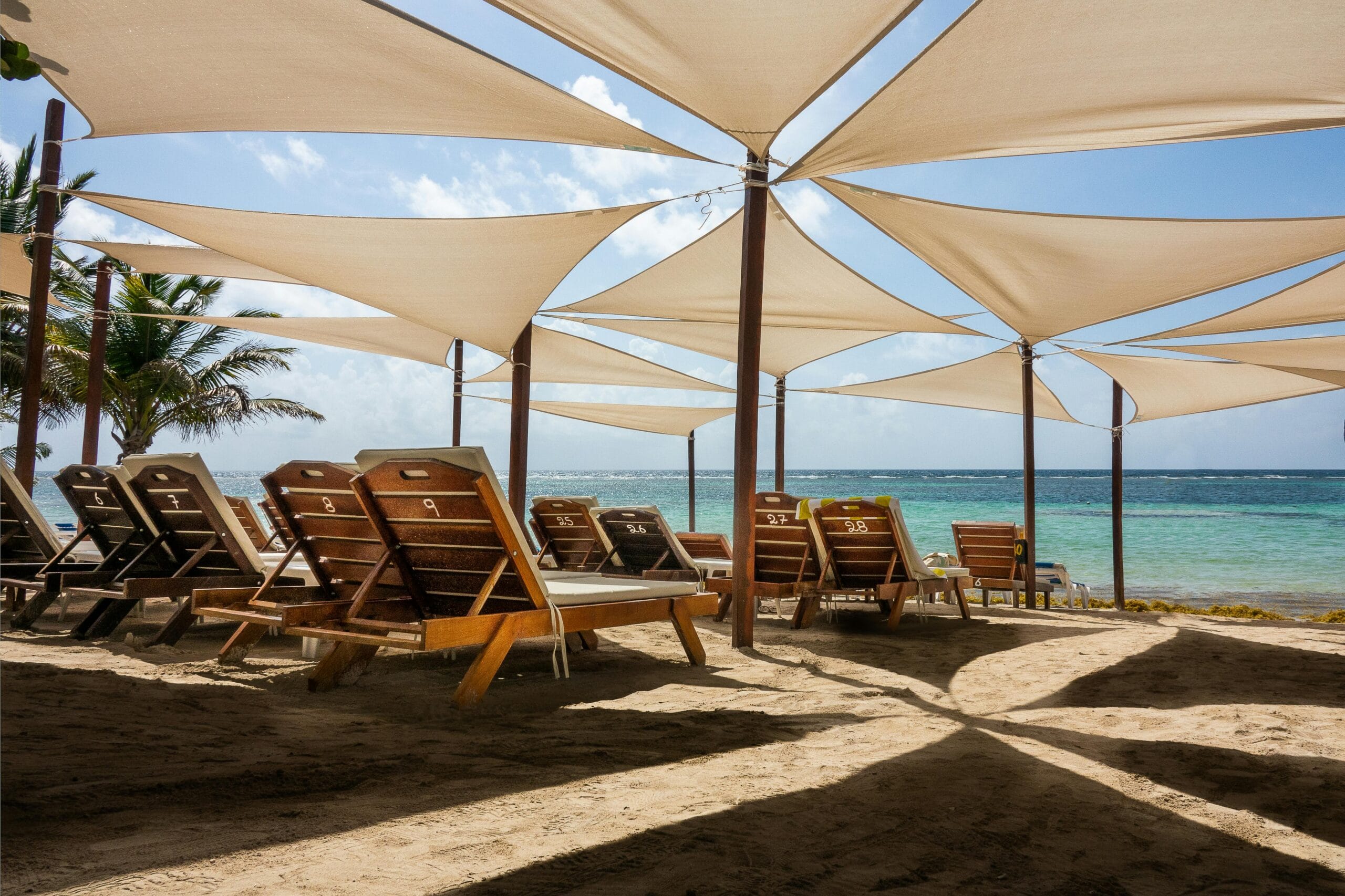 A serene beach scene featuring sun loungers under stylish shade sails by clear blue water.