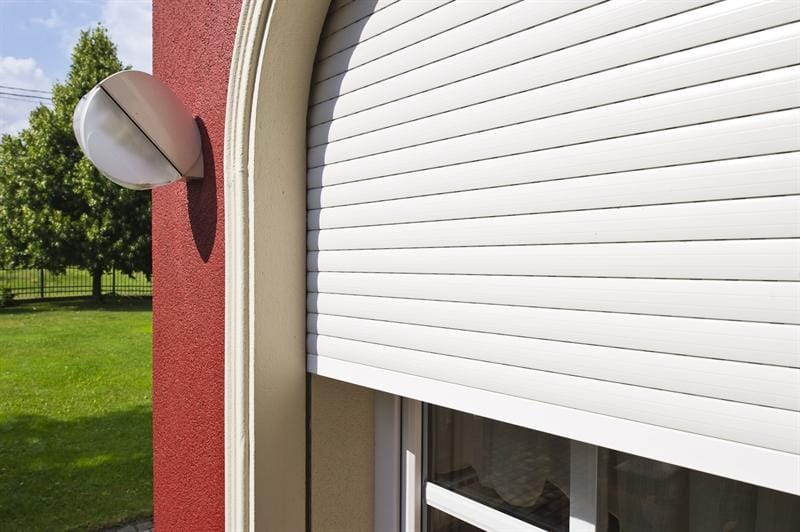Partial view of a white rolling shutter covering a window on a red wall, with an exterior light fixture and a grassy yard visible nearby.