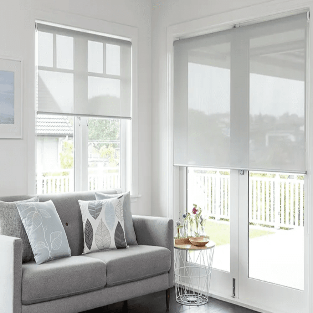 Bright living room with a gray sofa and decorative leaf-pattern cushions, near large windows with white roller blinds and a glass door to the outside side table with flowers nearby.