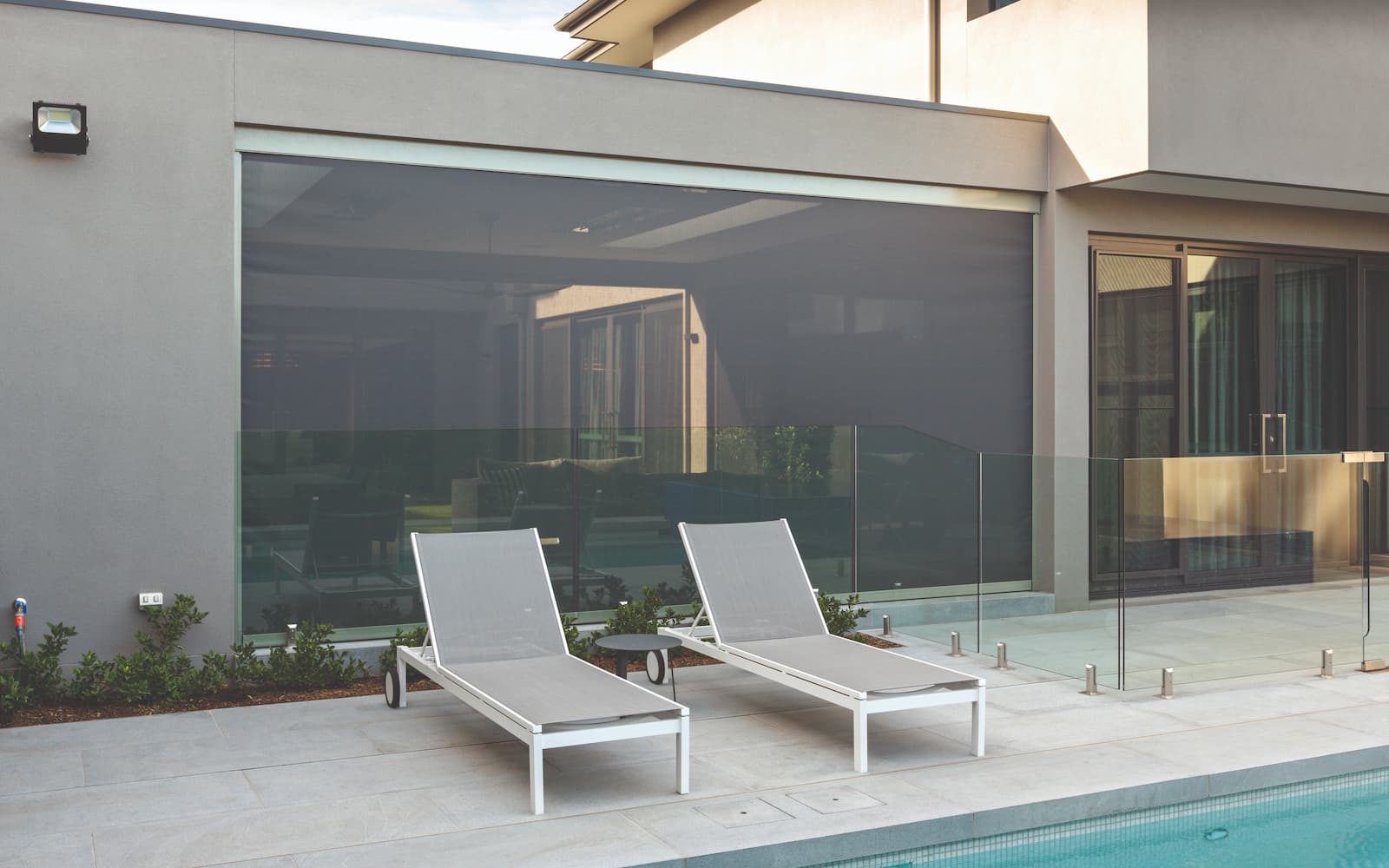 Two gray lounge chairs by a glass pool fence on a modern patio area, with a pool edge nearby.