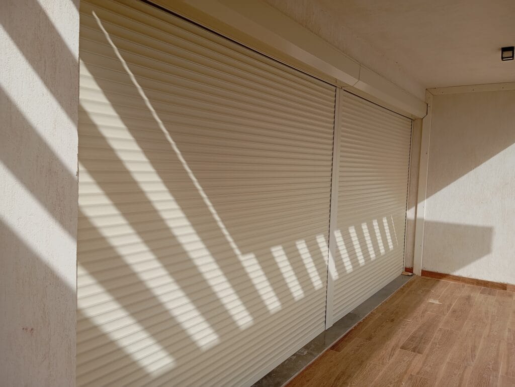 Closed beige rolling shutters along a hallway wall with diagonal shadows on the wall and wood floor nearby.