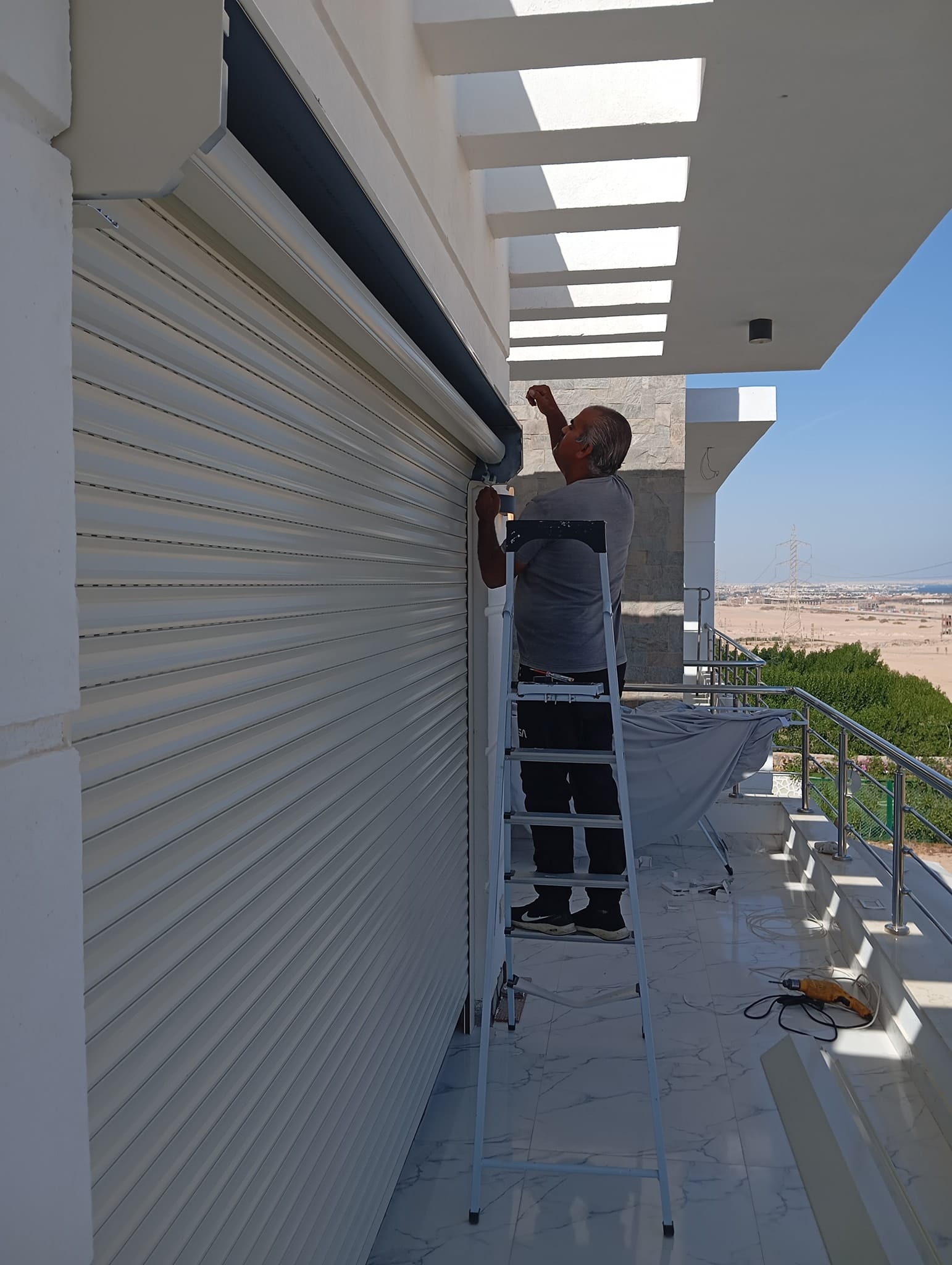 Man on a stepladder installing or adjusting a white rolling shutter on the building’s exterior, under a bright sunny sky.