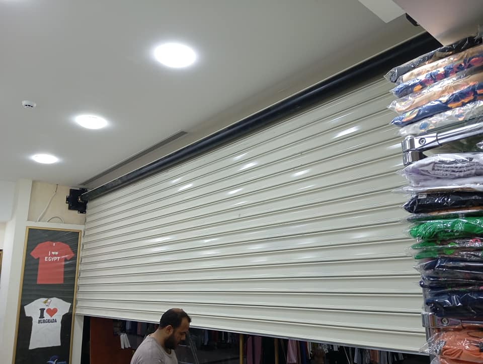 Store interior with a white rolling shutter down, bright ceiling lights, and racks of folded clothes; a man stands at the front left edge of the scene.
