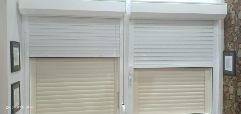 Two white-framed windows with beige roller shutters lowered, shown in a showroom setting with stone veneer on the right and framed documents on the sides.